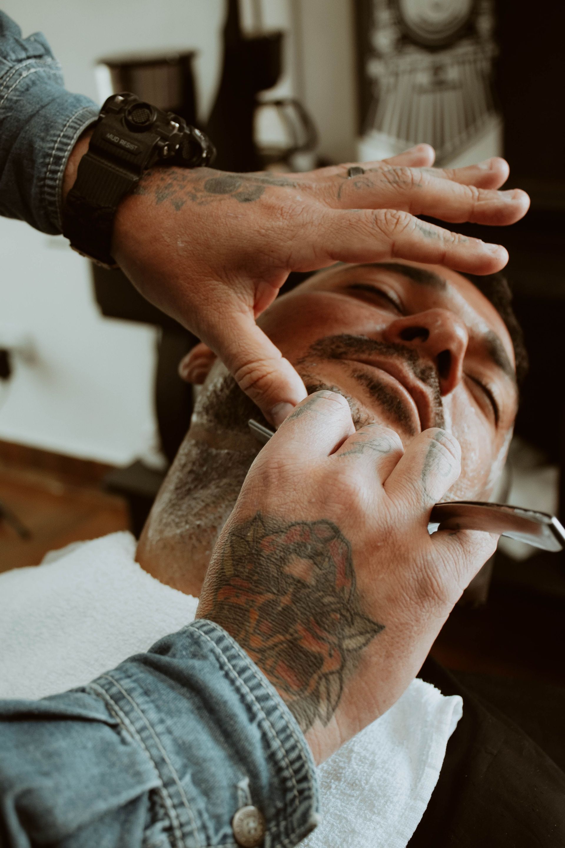 Close-up of a tattooed barber skillfully shaving a client's face in a modern barbershop.